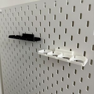 K&ouml;nnte beinhalten: Eine wei&szlig;e Lochwand mit gleichm&auml;&szlig;ig verteilten L&ouml;chern, mit zwei angebrachten Regalzubeh&ouml;rteilen. Ein Regal ist schwarz, das andere wei&szlig;. Die Regale haben mehrere F&auml;cher zur Aufbewahrung.