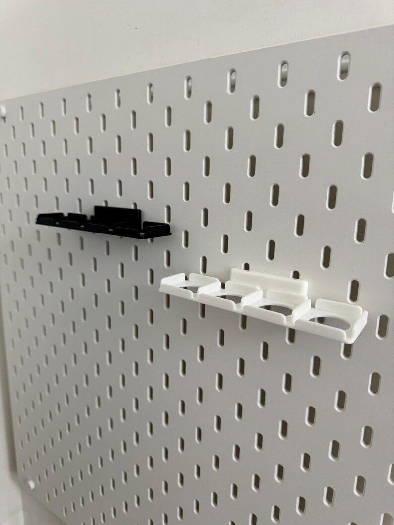 K&ouml;nnte beinhalten: Eine wei&szlig;e Lochwand mit gleichm&auml;&szlig;ig verteilten L&ouml;chern, mit zwei angebrachten Regalzubeh&ouml;rteilen. Ein Regal ist schwarz, das andere wei&szlig;. Die Regale haben mehrere F&auml;cher zur Aufbewahrung.