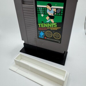 K&ouml;nnte beinhalten: Graue Nintendo Entertainment System-Spielkassette mit der Aufschrift "Tennis" und einer pixeligen Tennisszene. Die Kassette befindet sich in einem schwarzen St&auml;nder mit einer wei&szlig;en Ablage darunter.