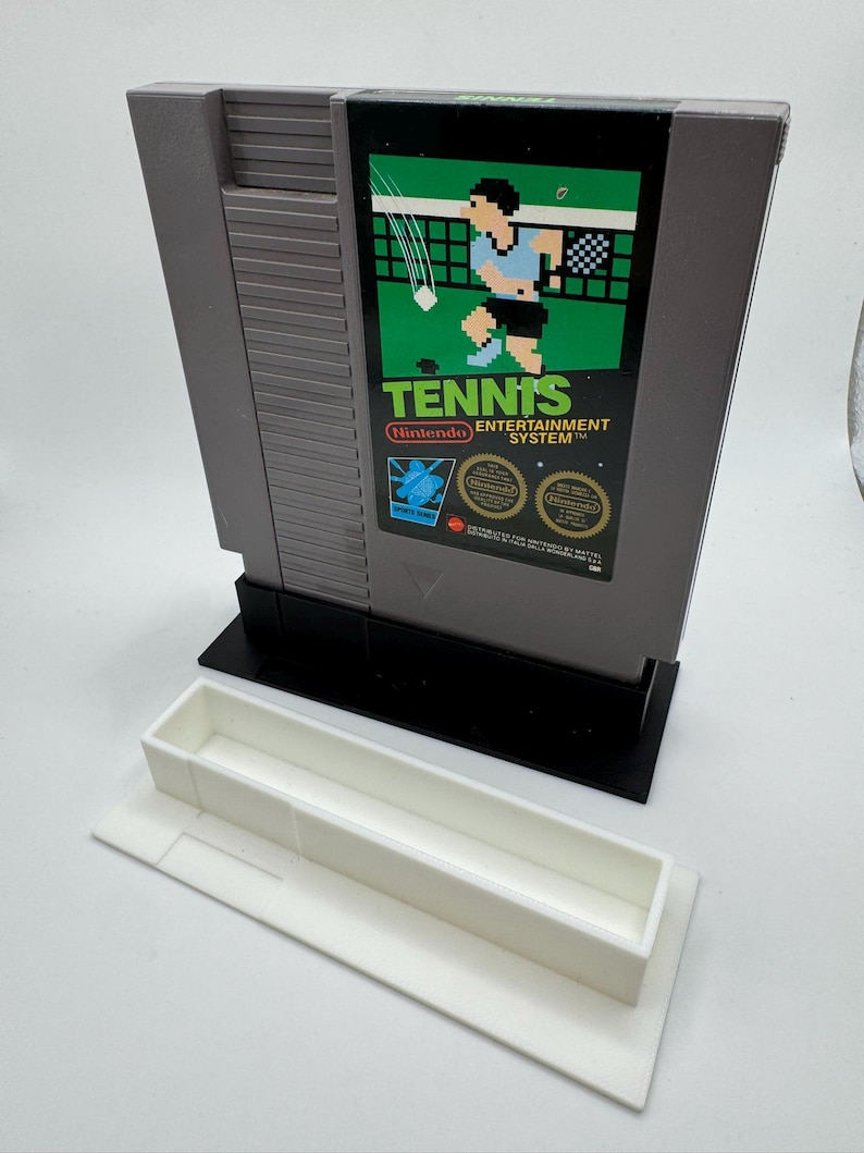 K&ouml;nnte beinhalten: Graue Nintendo Entertainment System-Spielkassette mit der Aufschrift "Tennis" und einer pixeligen Tennisszene. Die Kassette befindet sich in einem schwarzen St&auml;nder mit einer wei&szlig;en Ablage darunter.