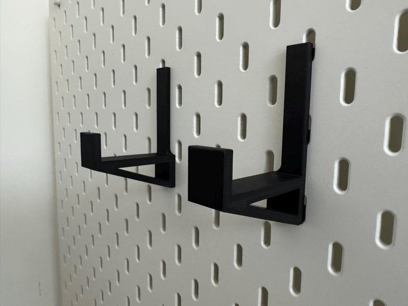 May include: Two black, L-shaped brackets designed for a pegboard. The brackets are made of a solid material and have a simple, functional design. They are mounted on a white pegboard with evenly spaced holes.