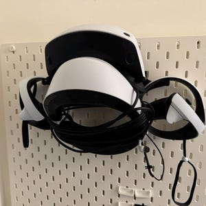 May include: White and black VR headset with attached controllers and cables, mounted on a white pegboard. The headset has a curved design with a black visor and a white body. The controllers are white with black accents.