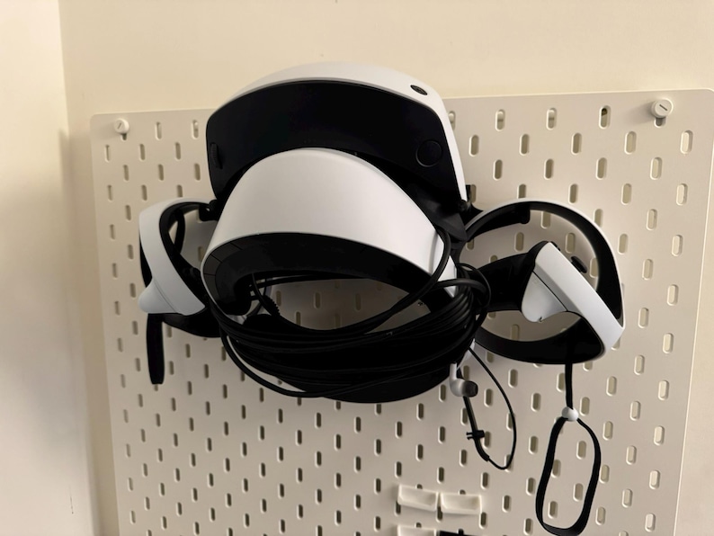 May include: White and black VR headset with attached controllers and cables, mounted on a white pegboard. The headset has a curved design with a black visor and a white body. The controllers are white with black accents.