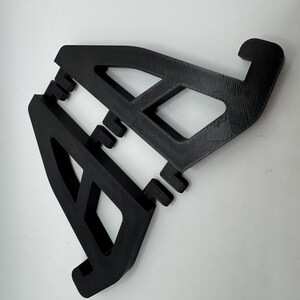 May include: Two black, 3D-printed phone stands. Each stand has an angled design with a rectangular cutout and a curved hook at the top. The stands are designed to hold a phone or tablet.
