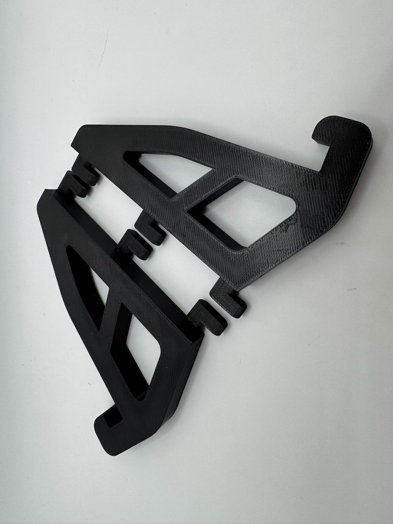 May include: Two black, 3D-printed phone stands. Each stand has an angled design with a rectangular cutout and a curved hook at the top. The stands are designed to hold a phone or tablet.