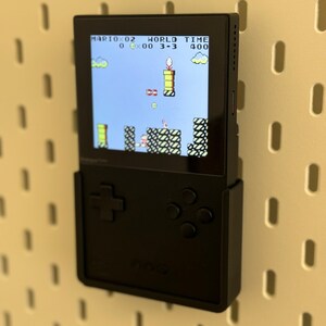 Op de afbeelding: Een zwarte draagbare videogameconsole met een scherm waarop het spel "Mario" wordt weergegeven met de tekst "MARIO 0-2 WORLD TIME 0 00 3-3 400". De console heeft een directionele pad en vier knoppen.