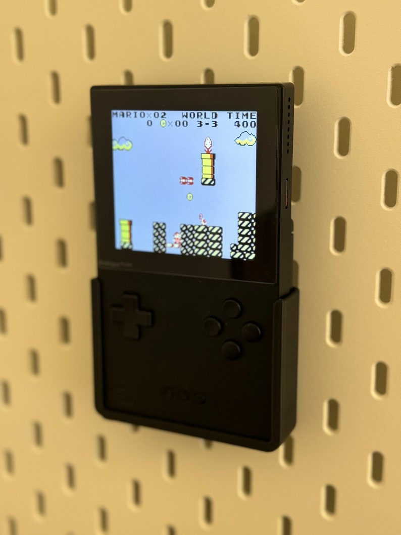 Op de afbeelding: Een zwarte draagbare videogameconsole met een scherm waarop het spel "Mario" wordt weergegeven met de tekst "MARIO 0-2 WORLD TIME 0 00 3-3 400". De console heeft een directionele pad en vier knoppen.