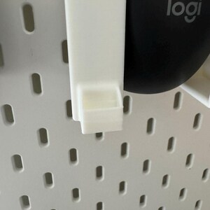 May include: A white plastic pegboard accessory with a small shelf. The accessory is designed to attach to a white pegboard with oval holes. A black computer mouse with the "logi" logo is visible in the background.
