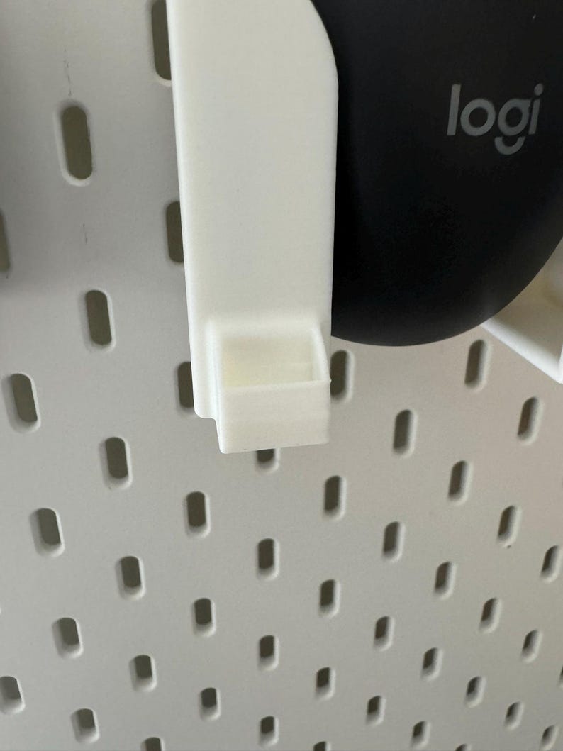 May include: A white plastic pegboard accessory with a small shelf. The accessory is designed to attach to a white pegboard with oval holes. A black computer mouse with the "logi" logo is visible in the background.