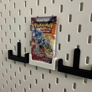 Op de afbeelding: Een Pok&eacute;mon-kaartspel boosterpack in een wit en oranje verpakking wordt weergegeven op een wit pegboard. Het pak wordt vastgehouden door een witte 3D-geprinte houder, geflankeerd door twee zwarte houders.