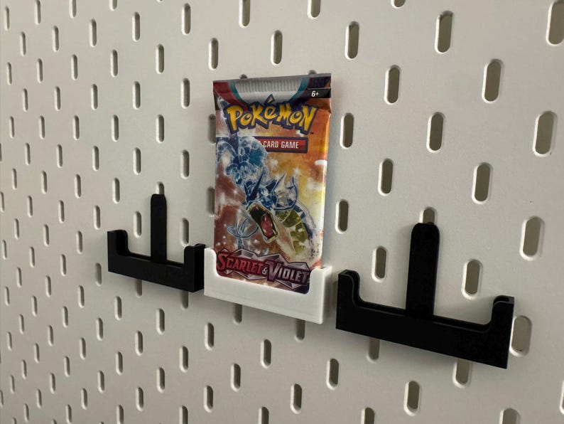 Op de afbeelding: Een Pok&eacute;mon-kaartspel boosterpack in een wit en oranje verpakking wordt weergegeven op een wit pegboard. Het pak wordt vastgehouden door een witte 3D-geprinte houder, geflankeerd door twee zwarte houders.