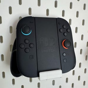 以下が含まれることがあります： 青とオレンジのアクセントが付いた黒いNintendo Switchコントローラー。 コントローラーにはボタンとジョイスティックがあり、白いペグボードに取り付けられています。 Nintendo Switchのロゴが見えます。