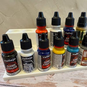 Puede incluir: Una colecci&oacute;n de botes de pintura Vallejo Game Color y Model Color en varios colores, incluyendo rojo, azul, amarillo y verde, expuestos en un soporte blanco escalonado. Los botes tienen tapas negras y est&aacute;n etiquetados con informaci&oacute;n del producto.