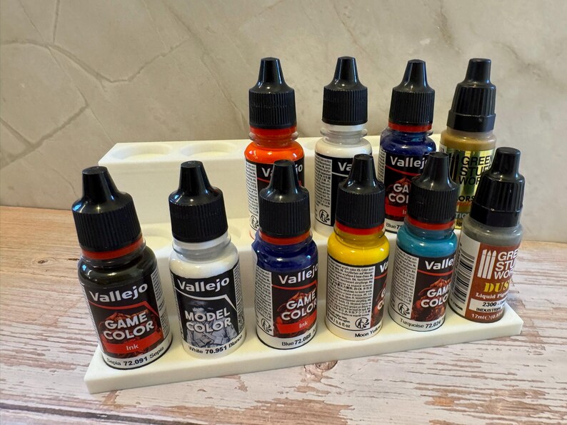 Puede incluir: Una colecci&oacute;n de botes de pintura Vallejo Game Color y Model Color en varios colores, incluyendo rojo, azul, amarillo y verde, expuestos en un soporte blanco escalonado. Los botes tienen tapas negras y est&aacute;n etiquetados con informaci&oacute;n del producto.