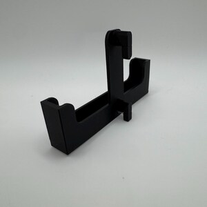 May include: A black, angled display stand with a rectangular base and a vertical support structure. The stand features a slot and a curved section, designed to hold an item. The stand is made of a solid, matte black material.