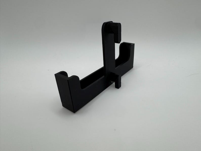 May include: A black, angled display stand with a rectangular base and a vertical support structure. The stand features a slot and a curved section, designed to hold an item. The stand is made of a solid, matte black material.