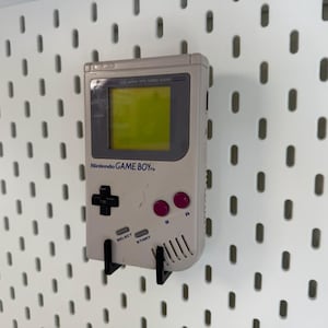 May include: A vintage Nintendo Game Boy handheld console in light gray, mounted on a white pegboard. The Game Boy features a green screen, black directional pad, pink buttons, and the text "Nintendo GAME BOY".