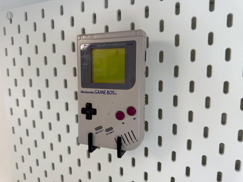 May include: A vintage Nintendo Game Boy handheld console in light gray, mounted on a white pegboard. The Game Boy features a green screen, black directional pad, pink buttons, and the text "Nintendo GAME BOY".