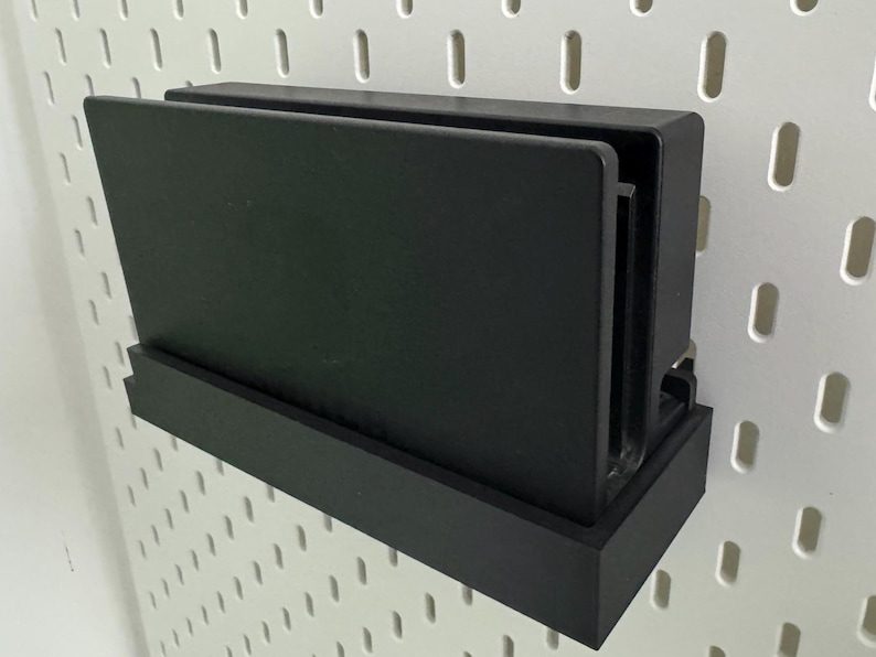 May include: A black wall-mounted storage shelf with a black rectangular base and a black rectangular top shelf. The shelf is designed to hold a gaming console.