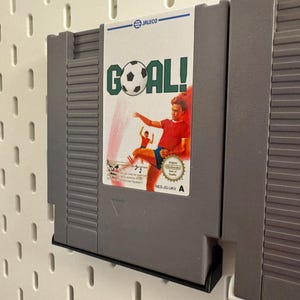 Pode incluir: Um cartucho de jogo cinza do Nintendo Entertainment System (NES) para o jogo "Goal!". O r&oacute;tulo apresenta uma bola de futebol e um jogador chutando a bola. O cartucho &eacute; montado em uma parede perfurada branca.