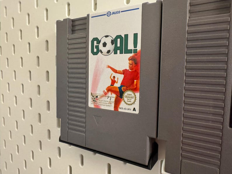 Pode incluir: Um cartucho de jogo cinza do Nintendo Entertainment System (NES) para o jogo "Goal!". O r&oacute;tulo apresenta uma bola de futebol e um jogador chutando a bola. O cartucho &eacute; montado em uma parede perfurada branca.