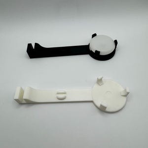 K&ouml;nnte beinhalten: Zwei 3D-gedruckte Handyst&auml;nder, einer schwarz und einer wei&szlig;. Beide St&auml;nder haben eine runde Basis mit einem erh&ouml;hten Rand und einen gebogenen Arm mit einem Haken am Ende.