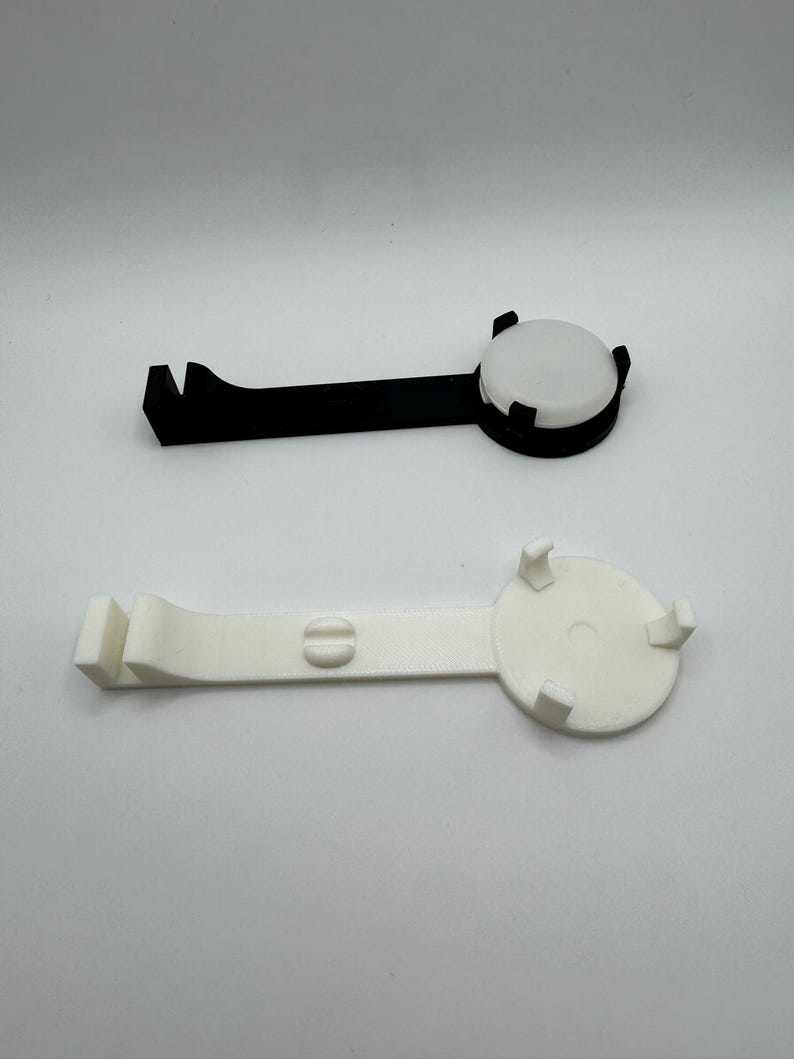 K&ouml;nnte beinhalten: Zwei 3D-gedruckte Handyst&auml;nder, einer schwarz und einer wei&szlig;. Beide St&auml;nder haben eine runde Basis mit einem erh&ouml;hten Rand und einen gebogenen Arm mit einem Haken am Ende.