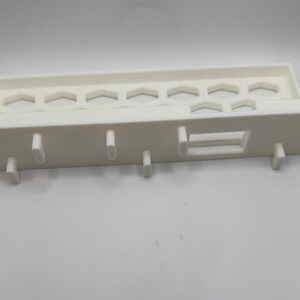 Puede incluir: Un estante de pl&aacute;stico blanco con un dise&ntilde;o rectangular. El estante tiene una serie de recortes hexagonales a lo largo de la parte superior y varias clavijas que sobresalen en la parte inferior. Tambi&eacute;n hay una abertura rectangular.