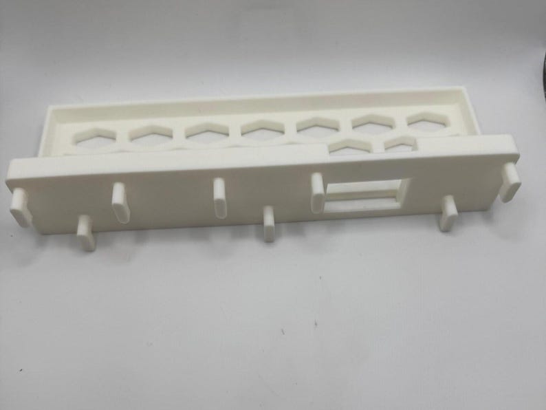 Puede incluir: Un estante de pl&aacute;stico blanco con un dise&ntilde;o rectangular. El estante tiene una serie de recortes hexagonales a lo largo de la parte superior y varias clavijas que sobresalen en la parte inferior. Tambi&eacute;n hay una abertura rectangular.