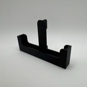 May include: A black plastic bracket with a T-shaped design. The bracket has a rectangular base with two raised sections on either end and a vertical support in the center. The object is set against a white background.