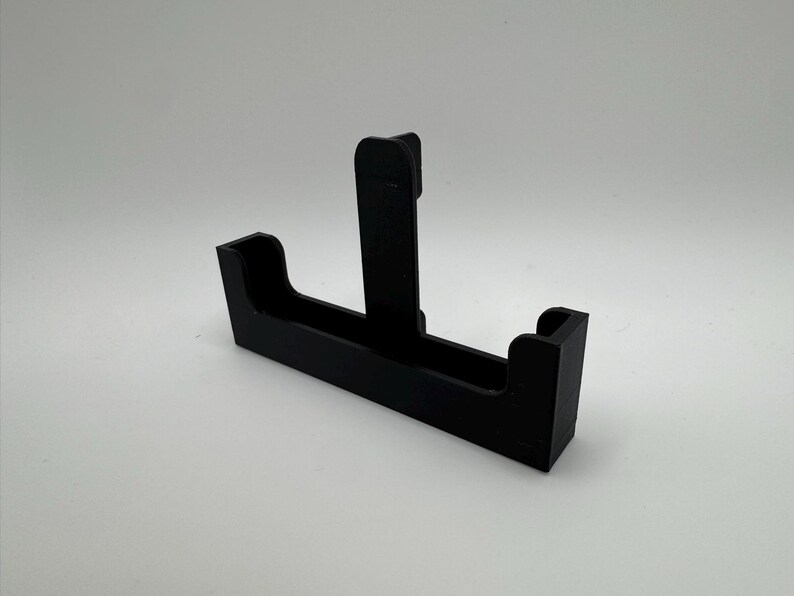 May include: A black plastic bracket with a T-shaped design. The bracket has a rectangular base with two raised sections on either end and a vertical support in the center. The object is set against a white background.