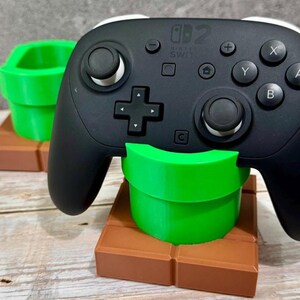 K&ouml;nnte beinhalten: Ein schwarzer Nintendo Switch Pro Controller liegt auf einem gr&uuml;nen, rohrf&ouml;rmigen St&auml;nder, mit einem zweiten St&auml;nder im Hintergrund. Die St&auml;nder stehen auf einer braunen Blockbasis. Der Controller hat wei&szlig;e Akzente und Tastenbeschriftungen.
