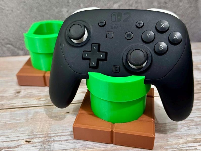 K&ouml;nnte beinhalten: Ein schwarzer Nintendo Switch Pro Controller liegt auf einem gr&uuml;nen, rohrf&ouml;rmigen St&auml;nder, mit einem zweiten St&auml;nder im Hintergrund. Die St&auml;nder stehen auf einer braunen Blockbasis. Der Controller hat wei&szlig;e Akzente und Tastenbeschriftungen.