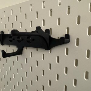 May include: A black pegboard hook is mounted on a white pegboard. The hook has a unique design with multiple arms and a curved hook. The pegboard has evenly spaced, oblong holes.