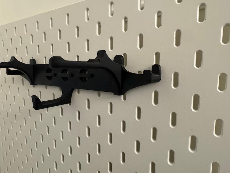 May include: A black pegboard hook is mounted on a white pegboard. The hook has a unique design with multiple arms and a curved hook. The pegboard has evenly spaced, oblong holes.