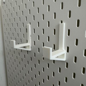 May include: Two white, 3D-printed pegboard hooks are mounted on a white pegboard. The hooks are L-shaped, designed to hold items. The pegboard has a grid of evenly spaced holes.