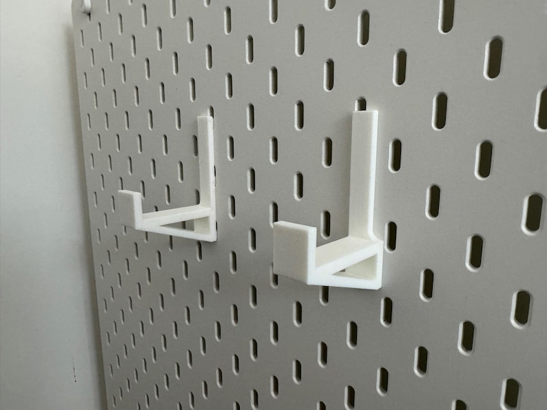 May include: Two white, 3D-printed pegboard hooks are mounted on a white pegboard. The hooks are L-shaped, designed to hold items. The pegboard has a grid of evenly spaced holes.