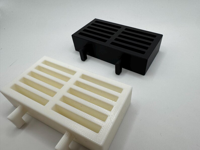 Op de afbeelding: Twee rechthoekige, sleufvormige, 3D-geprinte objecten. E&eacute;n is zwart, de andere is gebroken wit. Beide hebben een raster van rechthoekige openingen en twee steunpoten. De objecten zijn waarschijnlijk componenten of onderdelen voor een groter systeem.