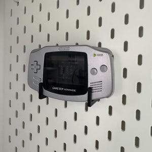 May include: A silver Game Boy Advance console mounted on a white pegboard. The screen shows a game, and the console has a directional pad, buttons, and the text "GAME BOY ADVANCE".
