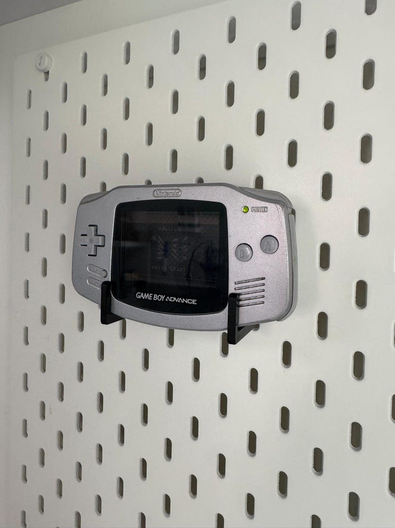 May include: A silver Game Boy Advance console mounted on a white pegboard. The screen shows a game, and the console has a directional pad, buttons, and the text "GAME BOY ADVANCE".