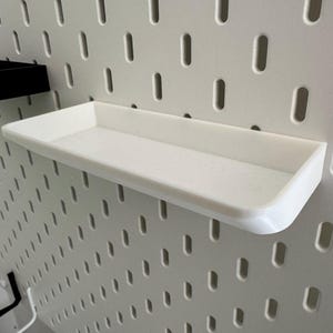 Puede incluir: Estante de pl&aacute;stico blanco con un borde elevado, montado en una pared de tablero de clavijas blanco.