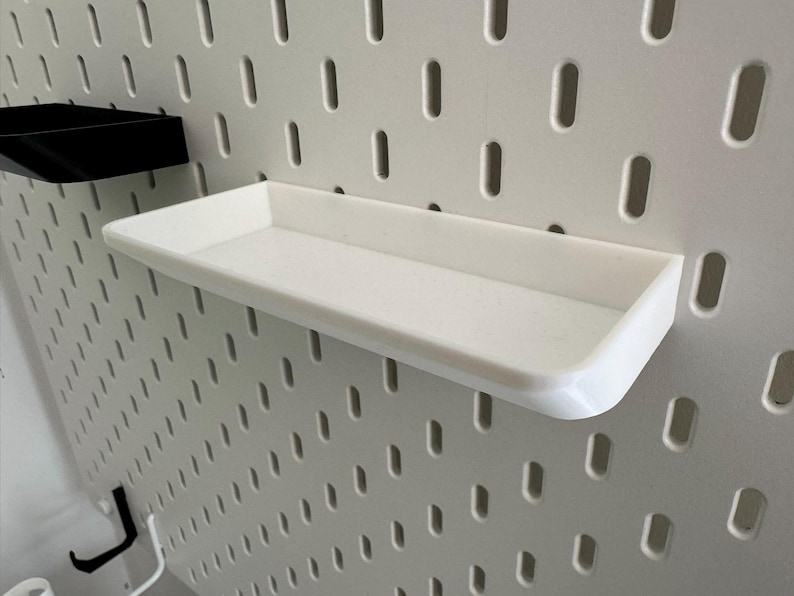 Puede incluir: Estante de pl&aacute;stico blanco con un borde elevado, montado en una pared de tablero de clavijas blanco.