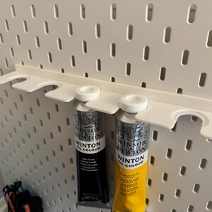May include: A white plastic pegboard shelf holding two tubes of Winsor & Newton Winton Oil Colour paint. One tube is black, the other is yellow. The pegboard has a series of evenly spaced oval holes.