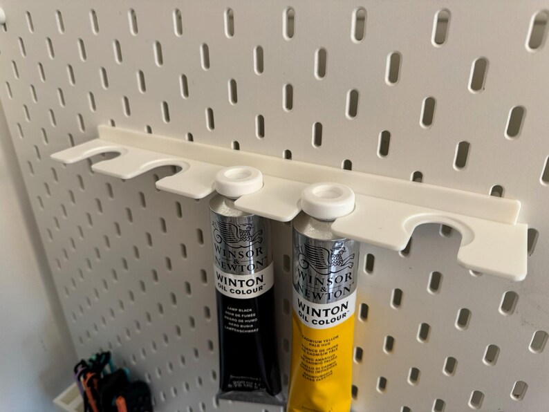 May include: A white plastic pegboard shelf holding two tubes of Winsor & Newton Winton Oil Colour paint. One tube is black, the other is yellow. The pegboard has a series of evenly spaced oval holes.