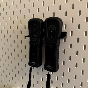 May include: Two black Wii remotes are mounted on a white pegboard. The remotes are in a holder, and each has a black wrist strap. The pegboard has a pattern of evenly spaced holes.