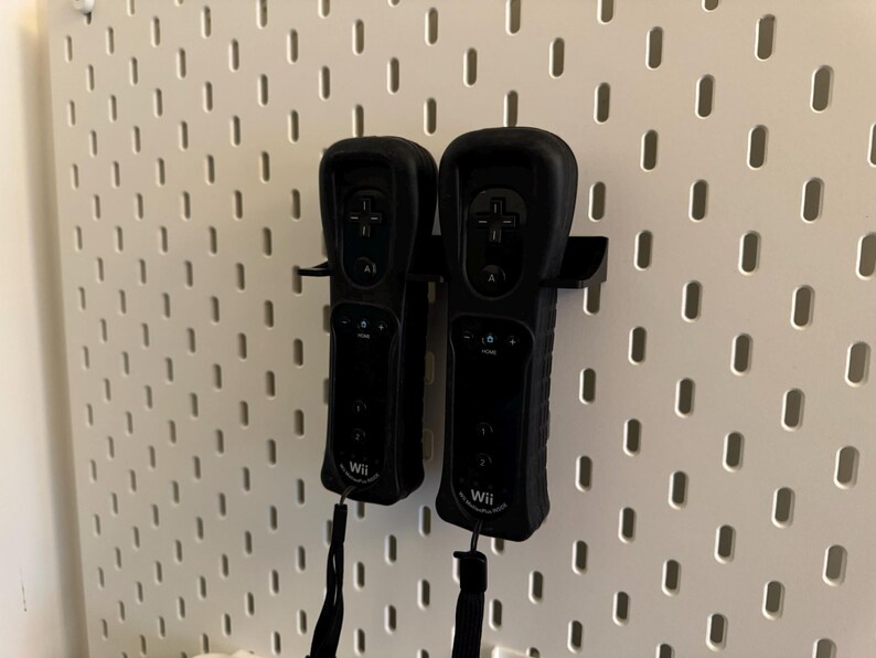May include: Two black Wii remotes are mounted on a white pegboard. The remotes are in a holder, and each has a black wrist strap. The pegboard has a pattern of evenly spaced holes.