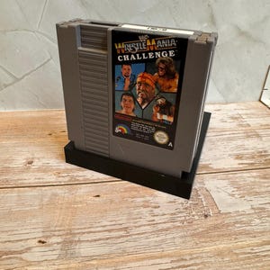 May include: A gray Nintendo Entertainment System (NES) game cartridge for WWF WrestleMania Challenge. The label features images of wrestlers and the game title. The cartridge is on a black stand, set against a light wood surface.