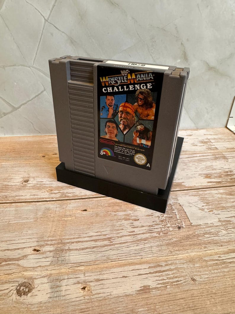 May include: A gray Nintendo Entertainment System (NES) game cartridge for WWF WrestleMania Challenge. The label features images of wrestlers and the game title. The cartridge is on a black stand, set against a light wood surface.