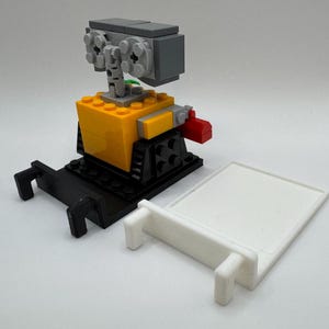 May include: A small, yellow and black robot made of Lego bricks. The robot has a gray head with a green plant growing out of it. The robot is standing on a black base with two black arms extending out. The robot is next to a white base with a small white arm extending out.
