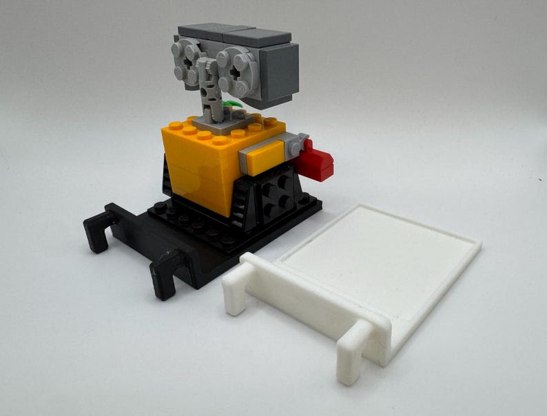 May include: A small, yellow and black robot made of Lego bricks. The robot has a gray head with a green plant growing out of it. The robot is standing on a black base with two black arms extending out. The robot is next to a white base with a small white arm extending out.